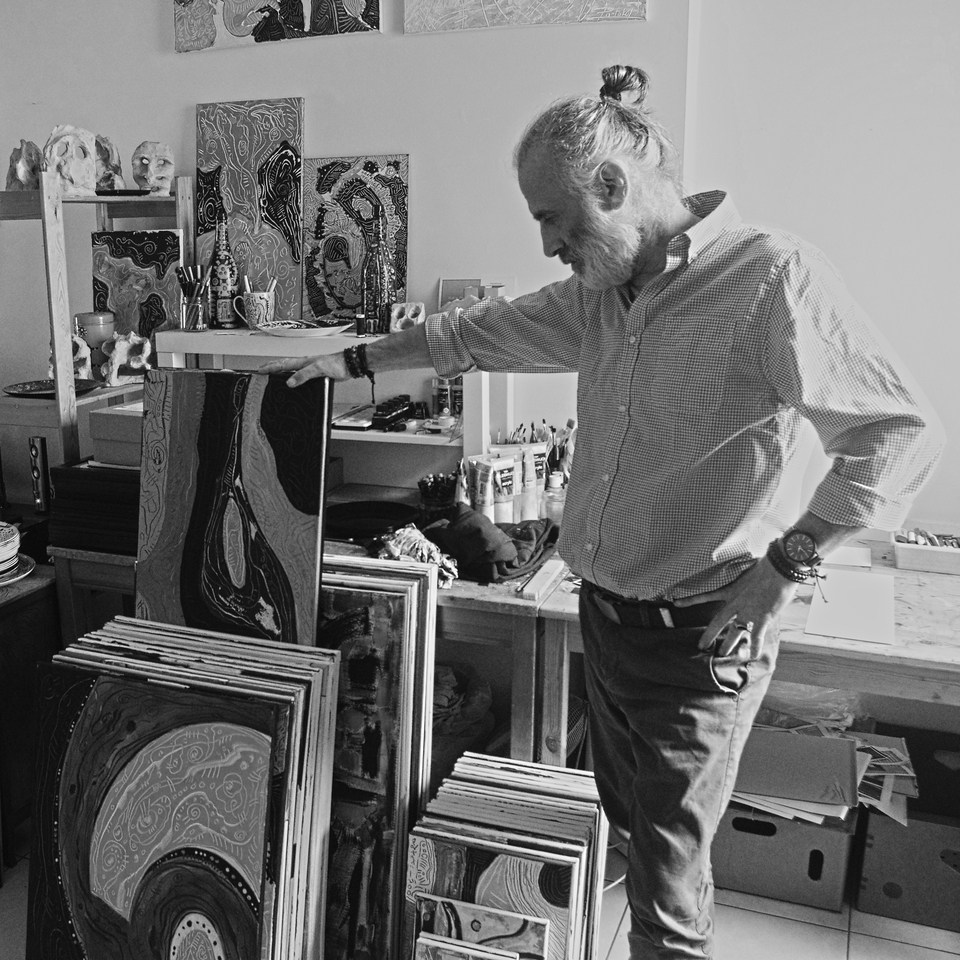 Dans l'atelier de l'artiste, devant les deux tables de travail à l'arrière-plan, à gauche de la photographie, se trouvent de nombreuses peintures abstraites. Hakan Portakal, en chemise, barbu, les cheveux attachés, la main droite sur l'un des tableaux, l'autre sur la hanche, dos à la lumière, regarde les tableaux. 