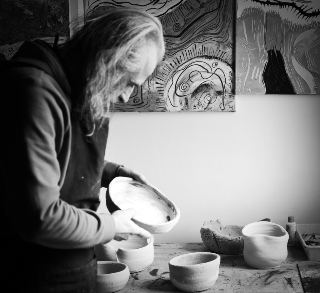 Hakan Portakal, en tablier de travail, à gauche, tourné vers la droite d’où elle vient la lumière. Il regarde une poterie à la main. Derrière lui sur la table, se trouve des poteries. Au mur on aperçoit deux tableaux abstraits. 