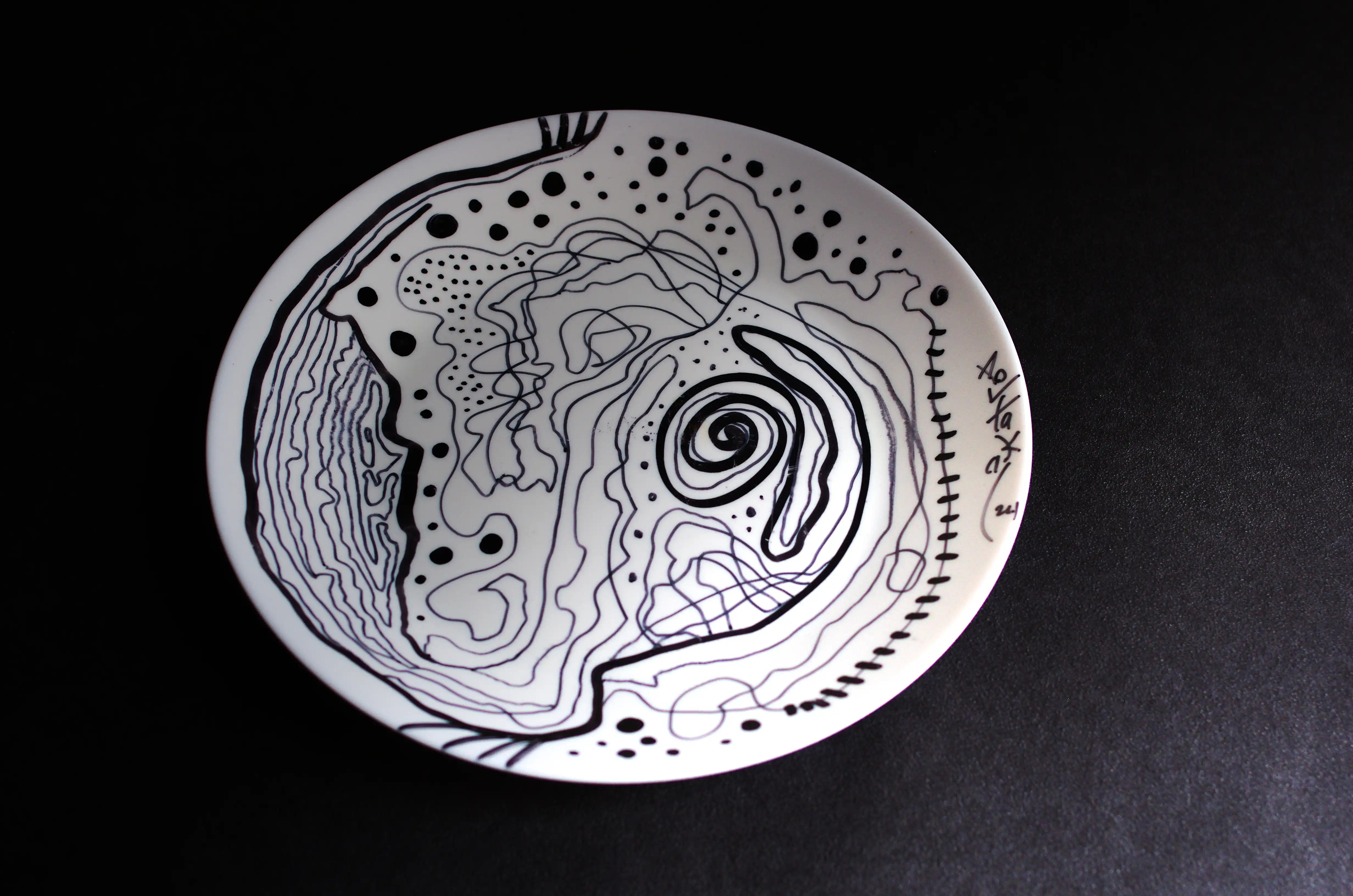 Cette assiette circulaire blanche est décorée de lignes noires courbes entrelacées. Ces lignes sont concentrées sur le côté gauche et l’une d’entre elles devient une spirale descendante et se prolonge vers l’extrémité droite de l’assiette. Il s’agit d’une œuvre d’art de qualité originale. Un choix d'investissement très esthétique. L'art abstrait n'a jamais été si élégant. Elle peut être achetée en toute sécurité. Abstrait | Brut | Original.