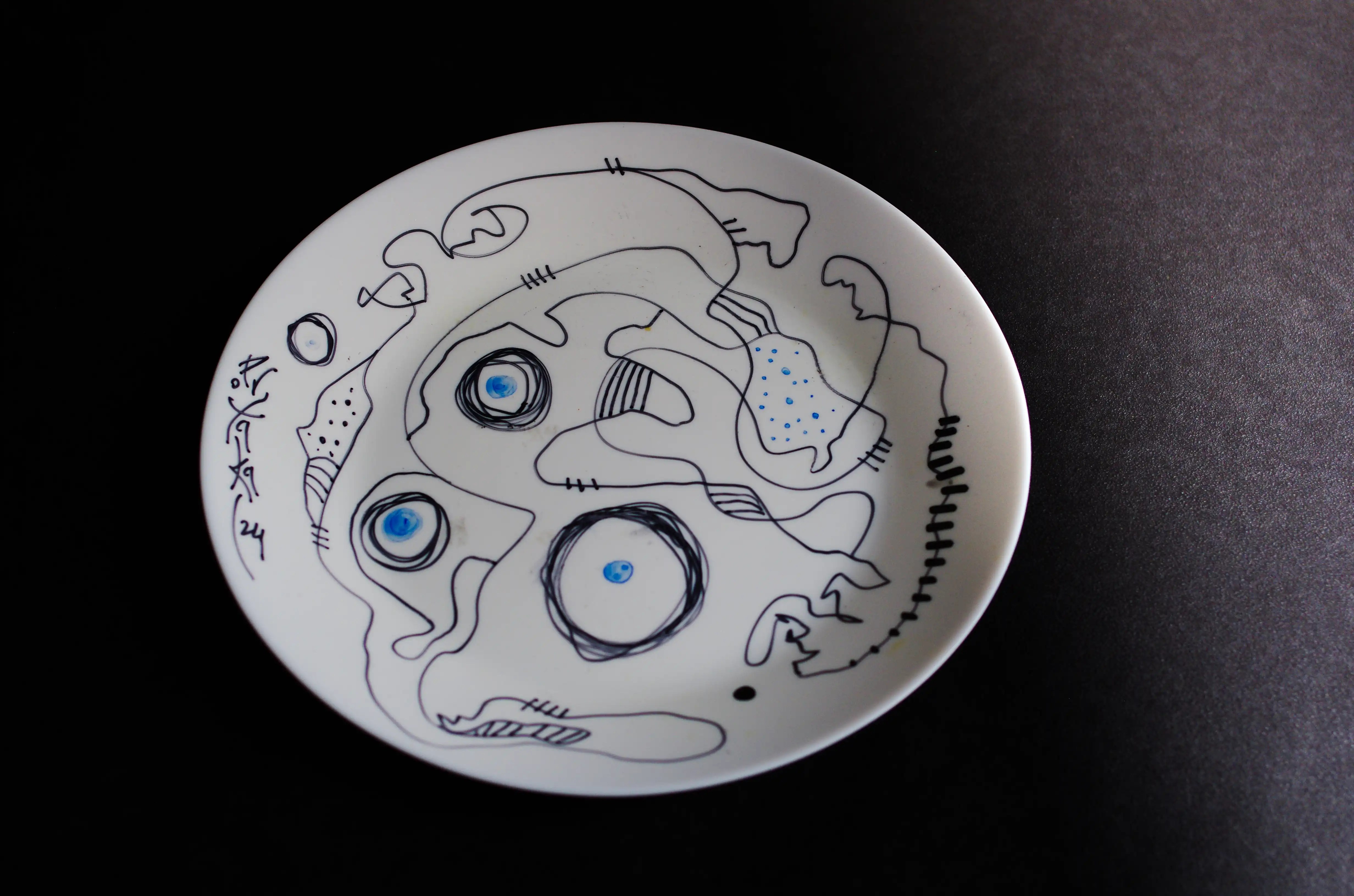 C’est une assiette circulaire blanche décorée de lignes noires courbes entrelacées. Il y a quatre anneaux faits de plusieurs lignes avec un point bleu au centre. Ces anneaux partent du côté gauche de l’assiette et se terminent en bas. Une belle pièce originale. Totalement subtil. Esthétisez fièrement vos investissements avec mes œuvres emblématiques.
