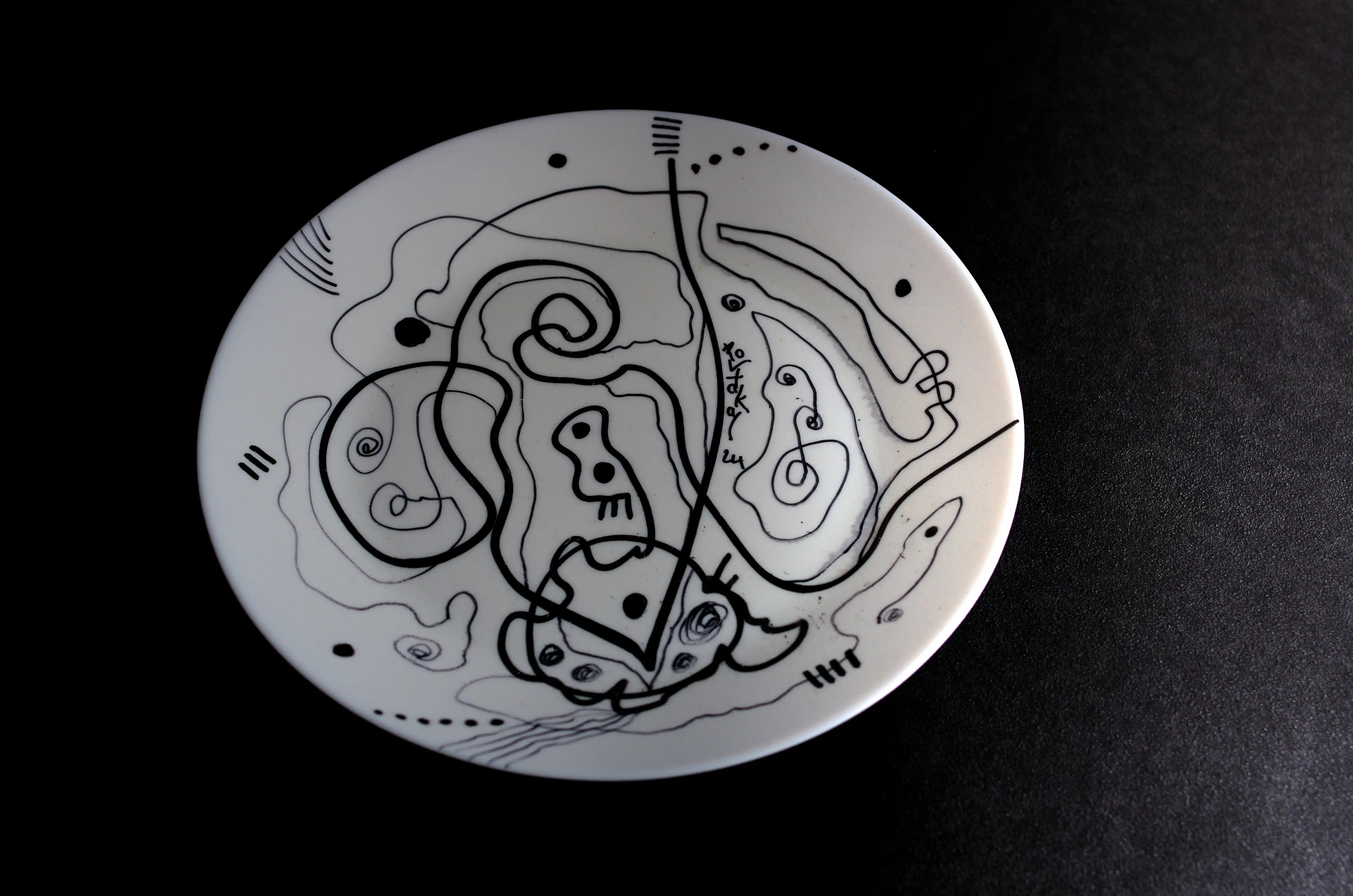 Une ligne noire épaisse, à partir des petites lignes horizontales en haut, descend vers le bas de l’assiette, fait un « V », puis un ovale et remontre vers le haut et devient une spirale. Une autre aussi  onduleuse  que la première départ de cette spirale et arrive le côté droit. Il y a un cercle au niveau de « V » qui l’enveloppe. Il y a également des lignes toutes fines, des points, etc, qui complètent la décoration de l’assiette. C’est une œuvre d’art originale, dynamique et de qualité. Achat en toute sécurité. Esthétisez vos investissements. Achetez maintenant, demain il sera peut-être trop tard. 