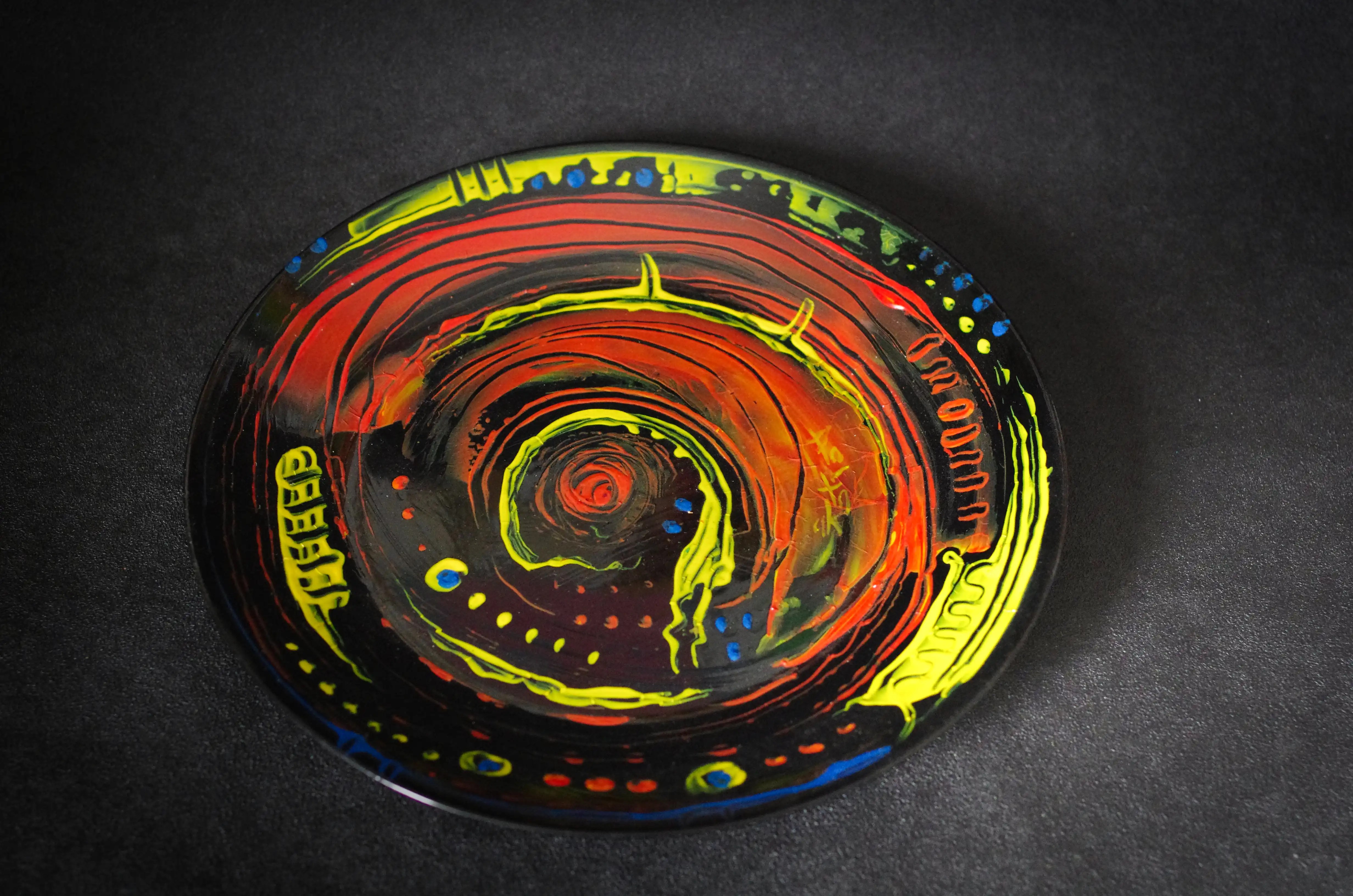 Noire à l'origine, cette assiette est magnifiquement décorée en acrylique jaune, rouge et bleu. Le mouvement commence au centre par un gros point rouge, suivi d'un anneau jaune entouré de rouge, puis à nouveau de jaune. La moitié du bord est jaune et décorée de petits points bleus, l'autre moitié est peu décorée. Achetez aujourd'hui pour gagner à l'avenir, en profitant de la beauté artistique. Faites honneur à votre portefeuille avec cette œuvre d'art extrêmement élégante et originale. Un achat judicieux est un investissement judicieux.