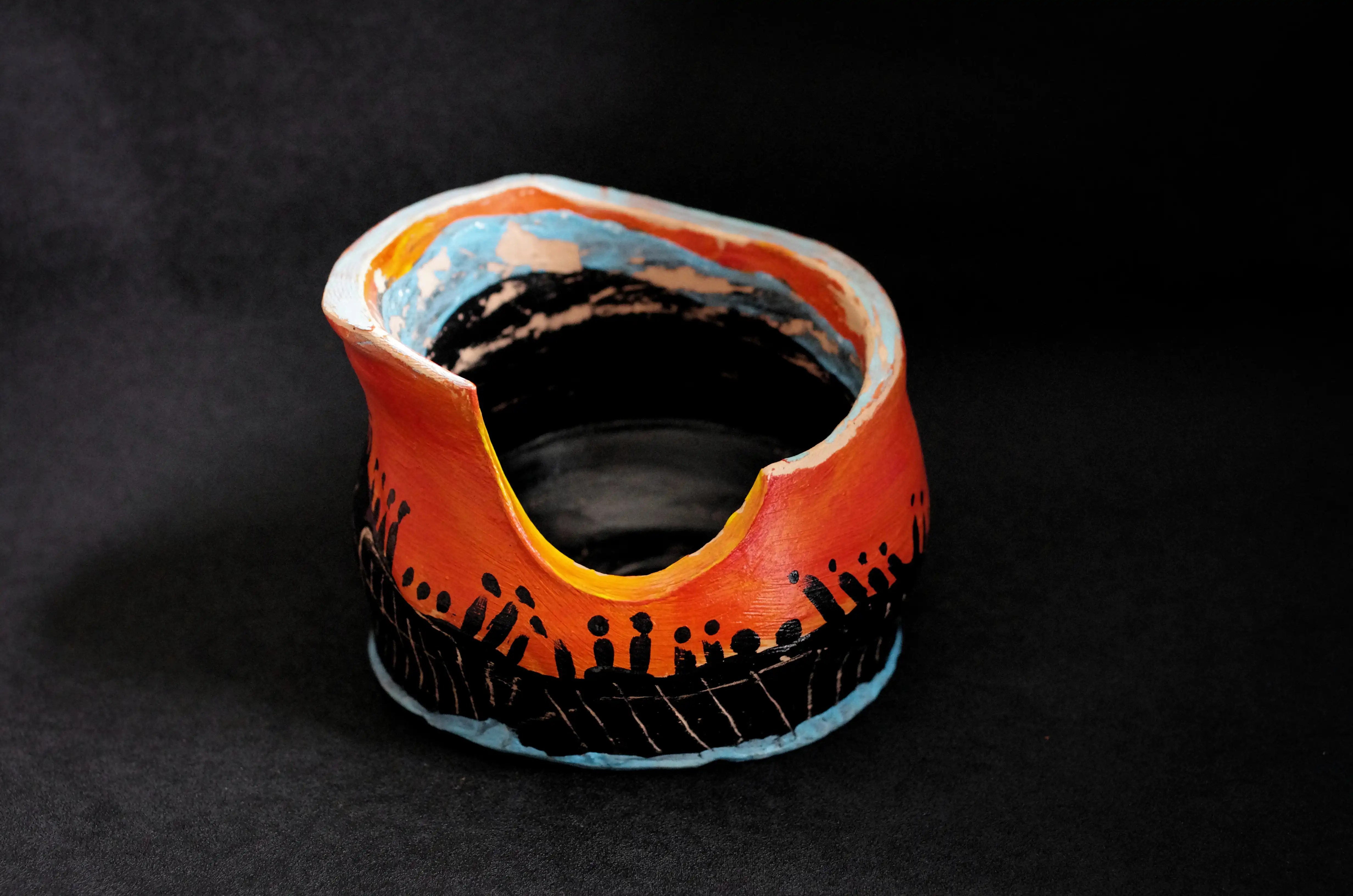 Cette poterie en argile cuite est une stylisation audacieuse dans la céramique artistique. C’est une poterie circulaire relativement haute et modelée d’une manière étonnante. Les bords sont taillés pour obtenir une surface plate. Il y a quatre bords dont trois sont bleus. Le quatrième est orange et le coupe descend quasiment jusqu’au bas. L’intérieur de cette œuvre est noir et près du bord, il y a une bande bleue et puis orange. À partir de quelques centimètres des bords à l’intérieur vers le fond, la peinture est grattée. Et à l’extérieur, cette poterie est, du bas vers le haut, bleue, noir et orange. La section noire et orange sont décorées. 