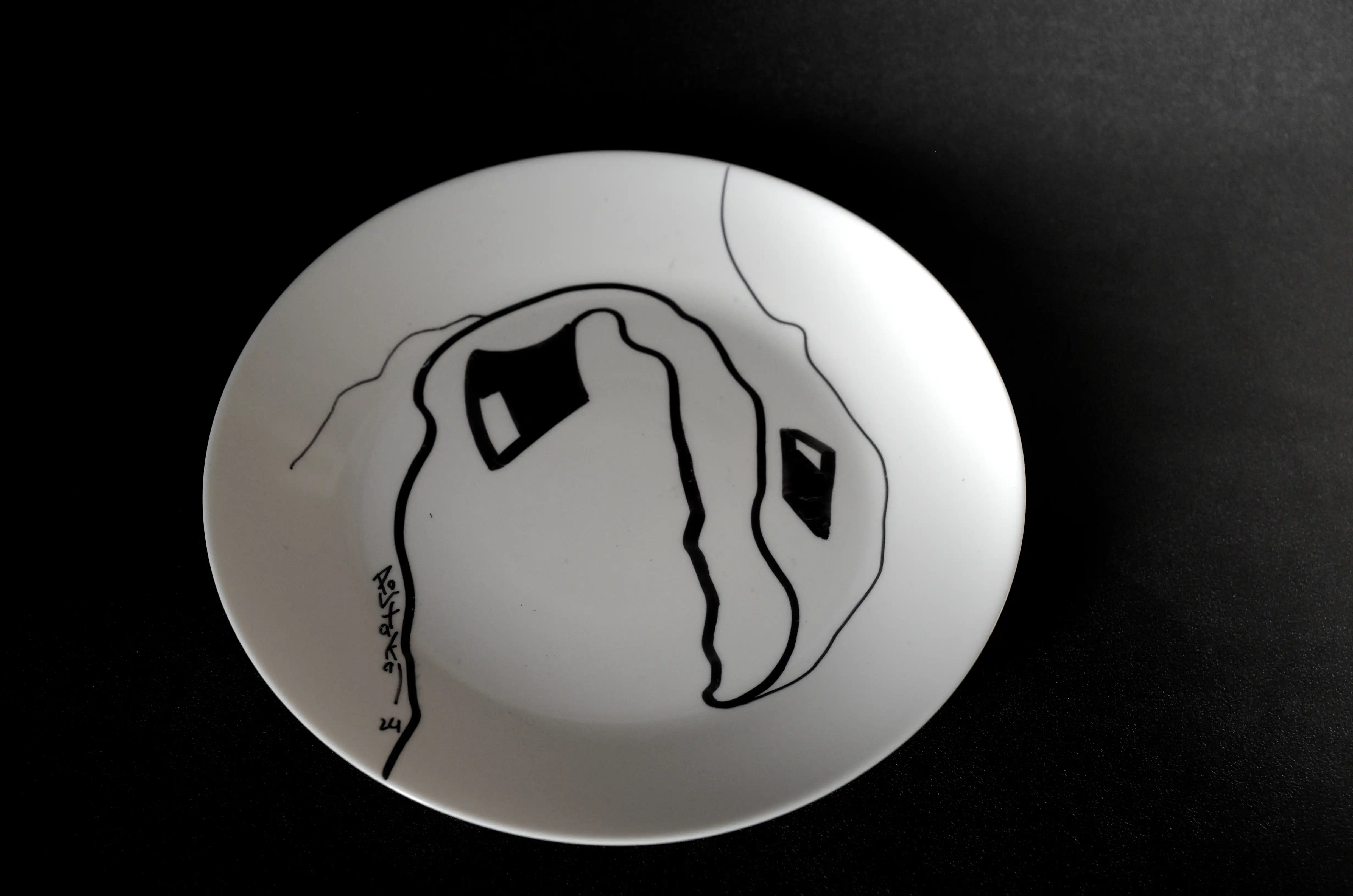 Il s'agit d'un ornement minimaliste réalisé au marqueur noir sur une assiette circulaire blanche. La composition consiste essentiellement en une ligne épaisse (un « U » renversé) qui s'étend vers le haut, vers le bas et vers le haut à partir de ma signature en bas à gauche. La ligne se termine par un carré déformé avec des espaces rectangulaires. Une ligne fine part du haut de la ligne épaisse vers la gauche de la plaque. Une autre ligne part du bas de la ligne épaisse vers le haut. Entre ces deux lignes se trouve un rectangle noir avec un espace identique.